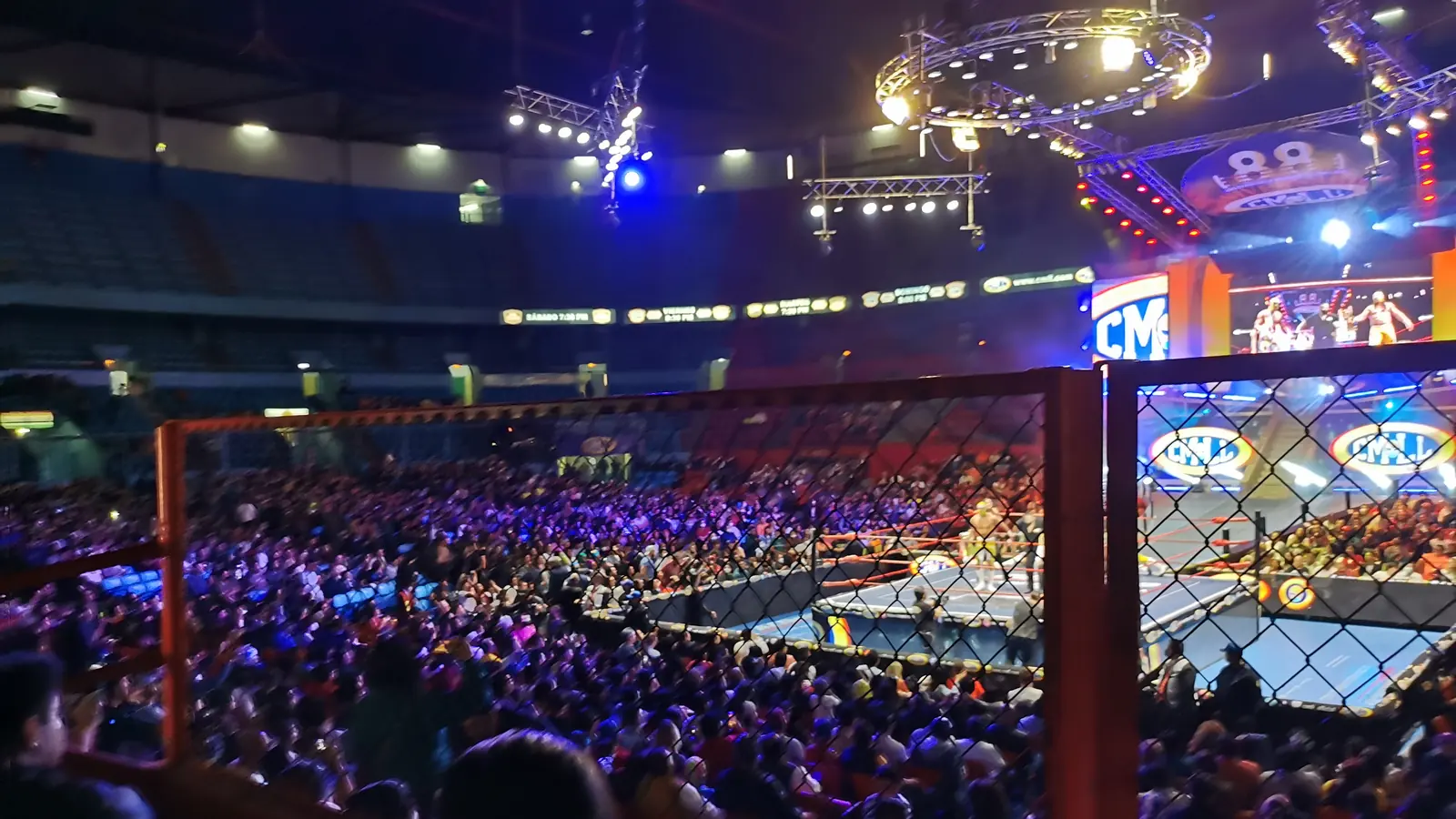 A crowd packs the stands of a Mexico City arena behind an orange safety fence, with the ring and big screens lit in blue and orange