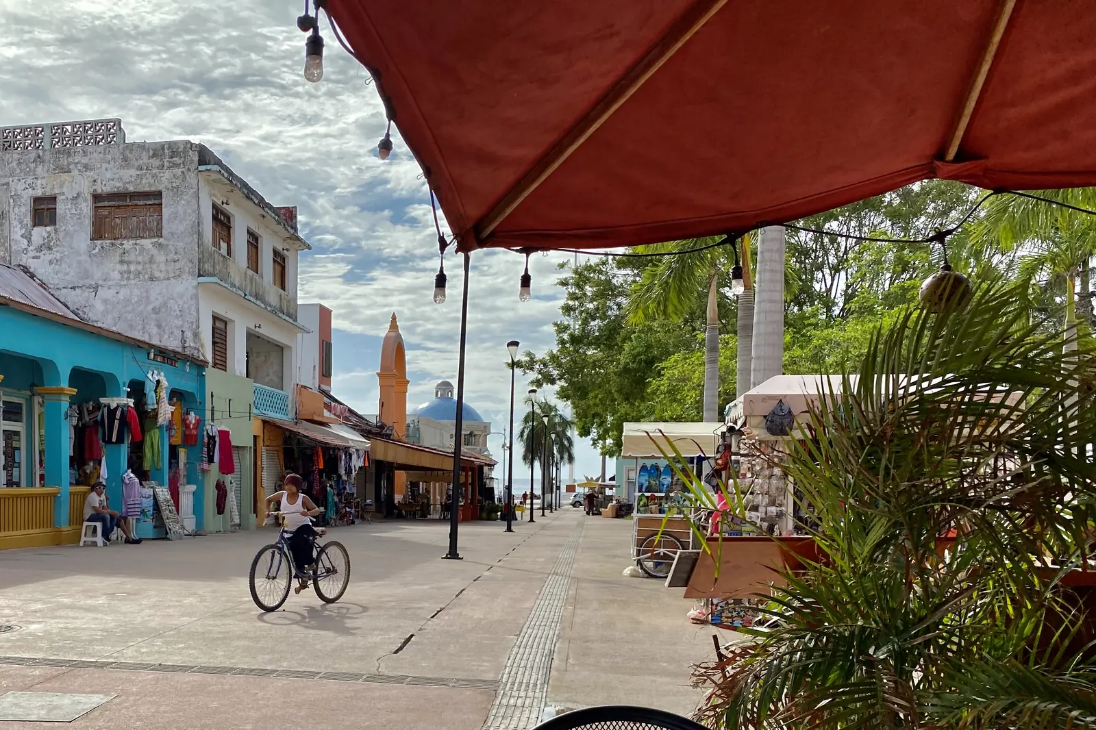 A cyclist rides along a San Miguel de Cozumel street of colourful shopfronts under an awning, with a church tower in the distance
